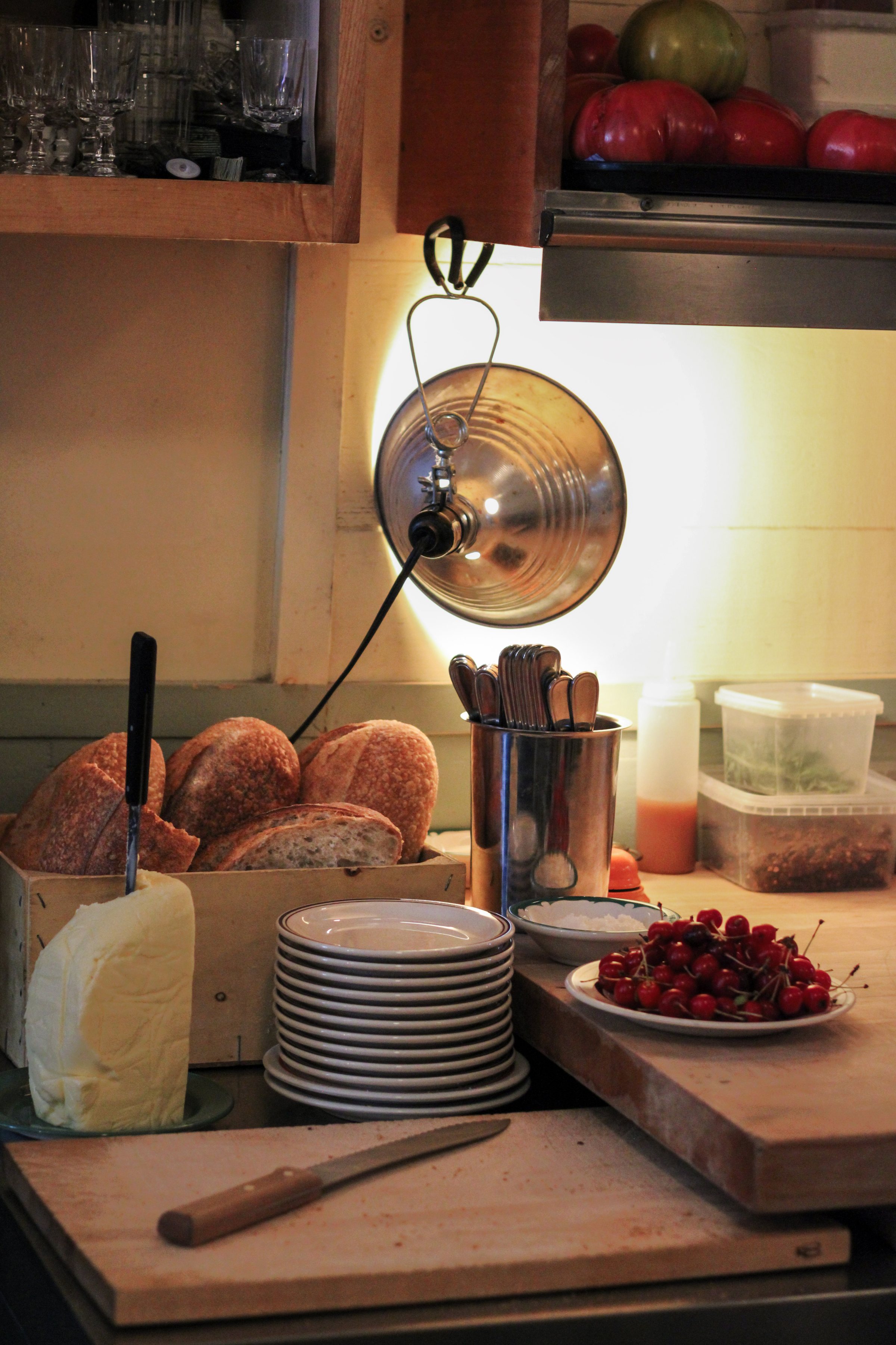 Prep station in a Montréal kitchen — bread, butter, cherries and stacked plates under a warm hanging lamp